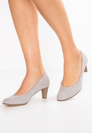 Femme portant des escarpins bas en suede gris clair avec des talons en bois empilés, se tenant sur un sol blanc.