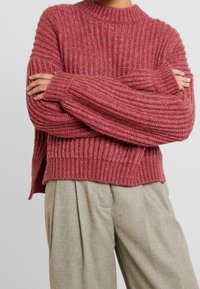 Pull en maille à large coupe dans un rose poussiéreux, avec des manches longues, un col haut et des fentes sur les côtés. Associé à un pantalon plissé gris clair.