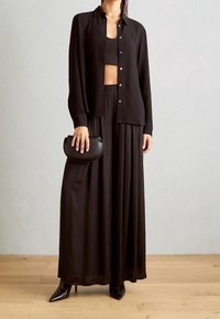 Camicia nera a maniche lunghe con bottoni sopra un top corto, abbinata a una maxi gonna plissettata. Tiene una piccola borsa tonda. Tacchi a spillo.