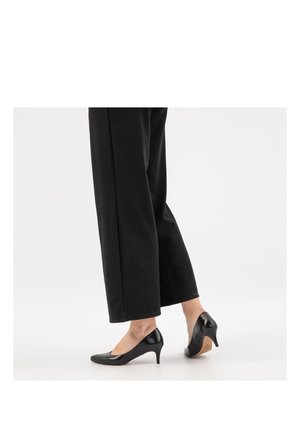 Personne portant un pantalon noir court et des escarpins en cuir noir à talon moyen, debout sur une surface blanche devant un fond uni.