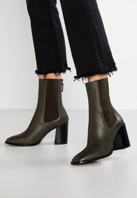 Zielone oliwkowe skórzane botki za kostkę z ostrym noskiem, elastycznymi bocznymi panelami i teksturowanym czarnym obcasem, z detalem zamka.