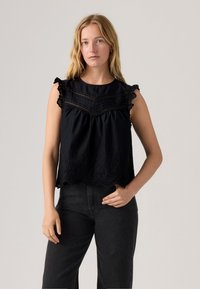 Top senza maniche nera con bordi scallopati, dettagli ricamati e accentuazioni con volant. Vestibilità ampia con arricciature sul petto.