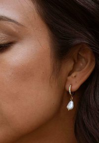 Goldene Hoops mit einem klaren Strassakzent und einer hängenden, unregelmäßig geformten Perle. Das Design ist elegant und minimalistisch.
