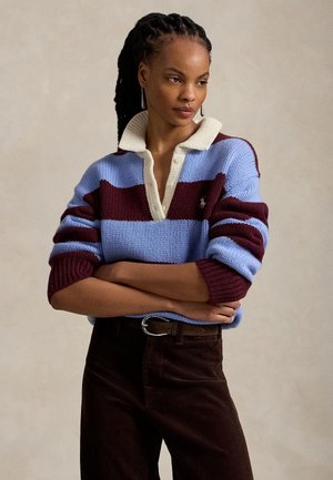 Femme aux cheveux tressés, vêtue d’un pull en tricot rayé bleu et bordeaux avec un col blanc, un pantalon marron et des boucles d’oreilles pendantes, bras croisés.