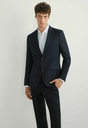 Młody mężczyzna o kręconych włosach, ubrany w dopasowany czarny garnitur i białą koszulę, stojący z jedną ręką w kieszeni na jednolitym tle.