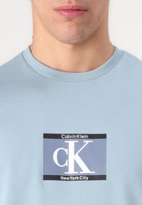 Lys blå bomull t-skjorte med et rektangulært svart og grått trykt logo med "Calvin Klein" og "New York City" i hvit tekst.