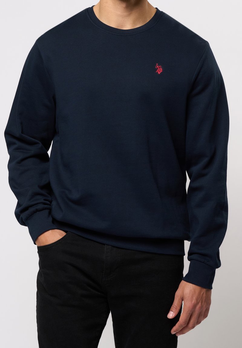 Homme portant un sweat-shirt à col rond à manches longues bleu marine avec un petit logo rouge brodé sur la poitrine gauche, associé à un pantalon noir.