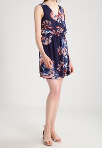Robe à imprimé floral en bleu marine avec un décolleté en V et une taille cintrée. Présente des fleurs colorées en rouge et rose. Tissu doux, design sans manches.