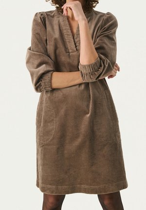 Persona con cabello rizado que lleva un vestido marrón de pana de manga larga con puños doblados, brazos cruzados, sobre un fondo blanco liso.