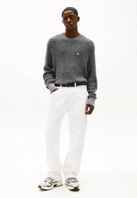 Maglione grigio lavorato a maglia con motivo testurizzato, jeans bianchi a gamba dritta e sneaker argentate, con un branding sottile sul maglione.