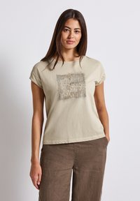 Béžové bavlněné tričko s krátkým rukávem, které má strukturovaný čtvercový design s textem "QUIET SENSES" v kontrastní barvě.