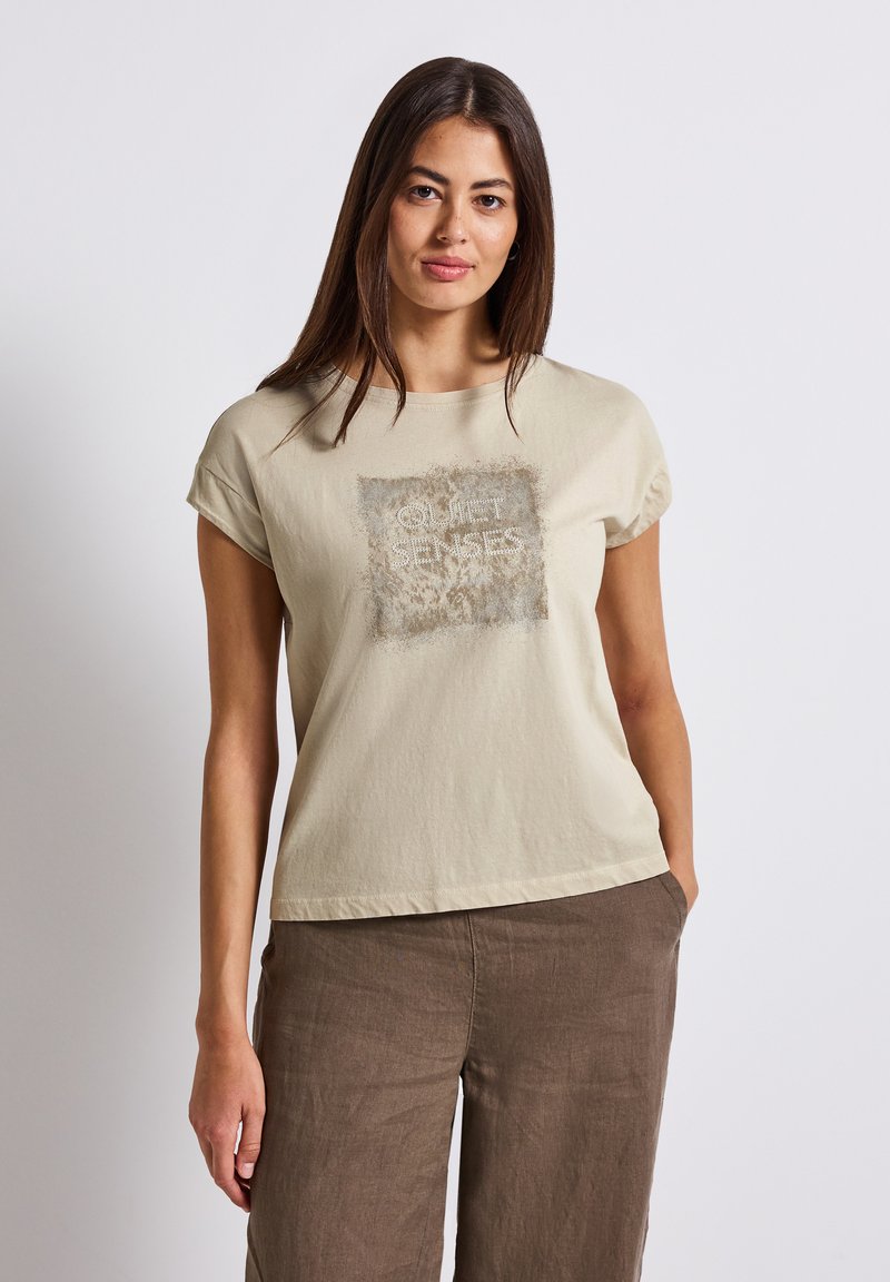 Béžové bavlněné tričko s krátkým rukávem, které má strukturovaný čtvercový design s textem "QUIET SENSES" v kontrastní barvě.