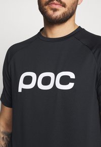 Černé sportovní tričko s krátkým rukávem, na kterém je vpředu velké bílé logo "poc", vyrobené z prodyšného materiálu s hladkou texturou.