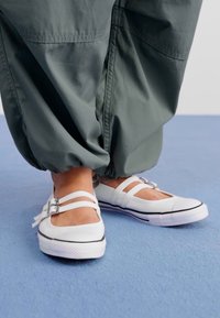 Pantofi plat albi cu două benzi și cataramă argintie, având vârful rotund, talpă albă cu margine neagră, purtați împreună cu pantaloni largi de culoare verde închis.