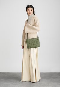 Zelená obdĺžniková taška s embosovaným vzorom, visiaca na tenkej šnúrke. Nosená s béžovým nadrozmerným svetrom a krémovými širokými nohavicami.