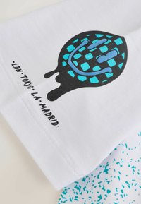 Weißes T-Shirt mit einem schwarz-blauen Grafikdesign, das ein stilisiertes Muster zeigt. Der Text lautet "LDN, TOKYO, LA, MADRID." Glatte Textur.