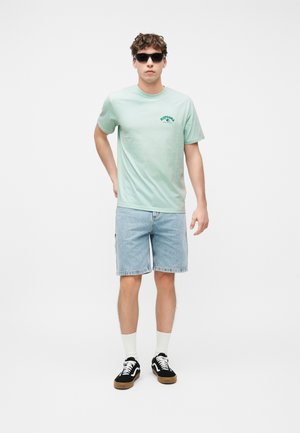 Bărbat purtând ochelari de soare negri, tricou verde deschis cu imprimeu, pantaloni scurți din denim albastru deschis, șosete albe și adidași negri, stând într-un decor cu fundal alb.