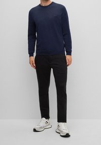 Pull bleu marine à col rond en tissu tricoté, associé à un pantalon noir et des chaussures de sport blanches, avec un logo discret sur la poitrine.
