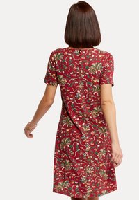 Vrouw met stijlbruin haar, gekleed in een rode jurk met korte mouwen tot de knie, versierd met groene, witte en gele bloemenprints, met haar rug naar de camera gekeerd.