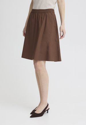 Mujer vestida con una falda marrón hasta la rodilla con cintura elástica y zapatos de tacón destalonados de punta oscura, de pie contra un fondo liso.