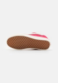 Baskets rouges et blanches couchées sur le côté, montrant une semelle en caoutchouc marron à motif gaufré sur un fond blanc uni.