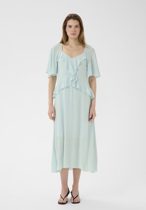 Frau trägt ein hellblaues Midikleid mit gestuften Rüschen und flatternden Ärmeln, kombiniert mit schwarzen Riemchensandalen, steht vor einem weißen Hintergrund.