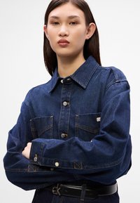 Δερμάτινο μπουφάν σε σκούρο μπλε χρώμα, με κουμπιά μπροστά, δύο τσέπες στο στήθος και κλασικό γιακά. Απολήγει με χρυσές ραφές και ασημένια κουμπιά.