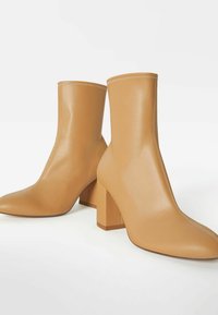 Par de botas de tobillo de cuero marrón con tacones de bloque y punteras afiladas, exhibidas sobre un fondo blanco.