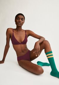 Burgunderfarbener Triangle-Bralette und passender Slip, kombiniert mit grünen Kniestrümpfen mit gelben Streifen, im Sitzpose vor einem hellen Hintergrund.