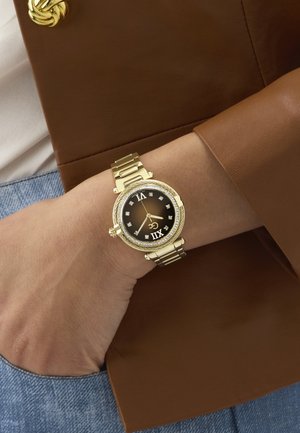 Polshorloge met een gouden metalen band en een zwarte wijzerplaat met Romeinse cijfers en kristallen markeringen, gedragen om de linkerpols over een bruine leren mouw.