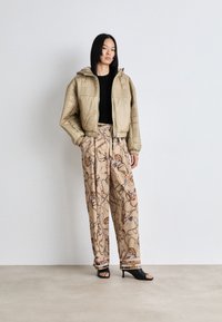 Beige quilted puffer jacket with a hood, paired with patterned beige trousers featuring chain and shoe designs, and black heeled sandals.
