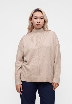 Jeune personne aux longs cheveux blonds portant un pull à col roulé en laine beige et un pantalon foncé, debout devant un fond clair uni.