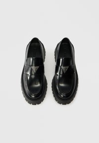 Black patent leather shoes with a chunky rubber sole, featuring a wide strap across the vamp and a triangular logo detail at the center.