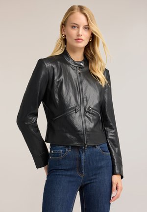 Frau mit langen blonden Haaren trägt eine schwarze Lederjacke mit Reißverschluss und blaue Jeans vor einem schlichten hellen Hintergrund.