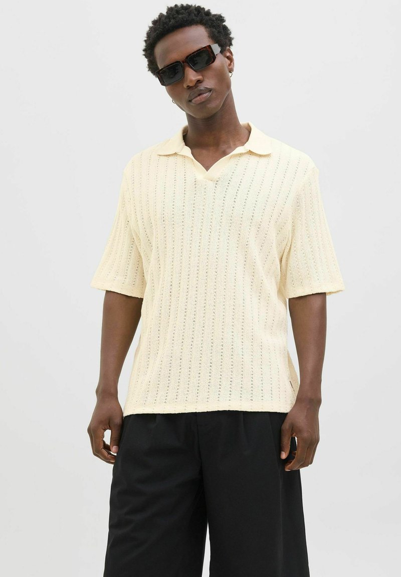 Polo shirt a maniche corte, di un giallo pallido, in maglia con scollo a V, che presenta costine verticali e una vestibilità ampia, abbinato a pantaloni neri a gamba larga.