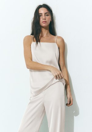 Žena s dugom tamnom kosom, odjevena u svijetlo bež top bez rukava i odgovarajuće hlače, stoji ispred jednobojne bijele pozadine.