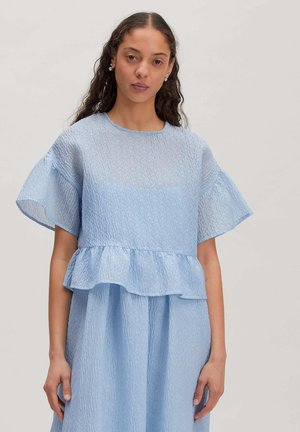 Frau mit langen lockigen Haaren, die eine hellblaue, strukturierte Kurzarmbluse mit Rüschensaum und einen passenden Rock vor einfarbigem Hintergrund trägt.