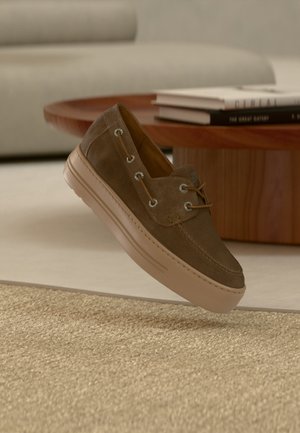 Chaussure décontractée en daim marron avec semelle en caoutchouc beige flottant au-dessus d'un tapis beige texturé, table basse en bois et piles de livres en arrière-plan.