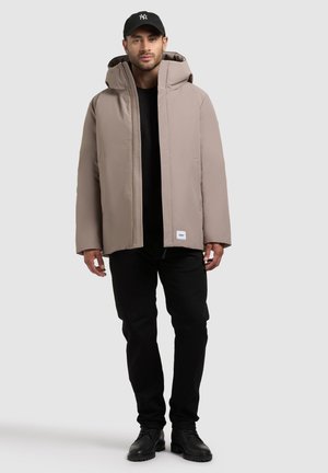 Giacca imbottita beige con un grande cappuccio, chiusura frontale con zip e bottoni a pressione. Indossata sopra una maglietta nera e abbinata a pantaloni e scarpe nere.