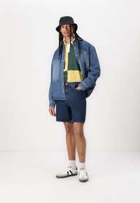 Denimjakke, grønn og gulstripete polo, blå denimshorts, svart bøttehatt, og hvite joggesko med svarte detaljer og grå sokker.