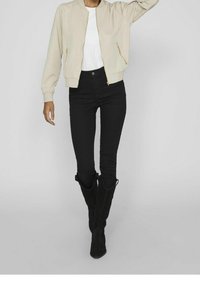 Creme bomberjakke med lynlåsdetaljer, parret med sorte skinny jeans og knæhøje sorte ruskindsstøvler. Modellen står med den ene hånd hævet.