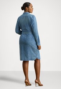 Žena stojaca z profilu, oblečená v rifľových šatách s opaskom a dlhými rukávmi, v hnedom sandále na vysokom opätku, na jednofarebnom pozadí.