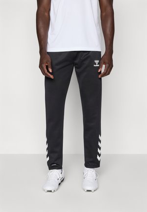 Mann trägt schwarze Sporthosen mit weißen Chevron-Streifen nahe den Knöcheln und weiße Sneaker, steht vor einem schlichten, hellen Hintergrund.