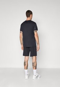 Μαύρο αθλητικό μπλουζάκι και σορτς, λείο ύφασμα, κοντά μανίκια, με λογότυπο στη μανσέτα, συνδυασμένο με λευκά αθλητικά παπούτσια και κοντές κάλτσες.