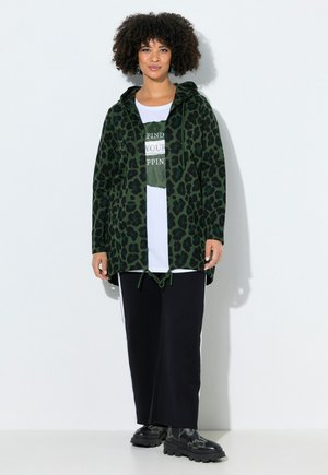 Kvinde med krøllet hår iført grøn leopardprint-hoodie, hvid graphic t-shirt, sorte vide bukser og sorte støvler, stående mod en hvid væg.