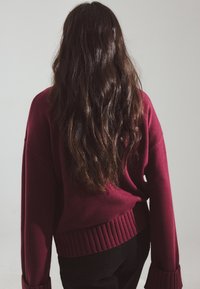 Burgundska pletena svetra z ribastim robom in manšetami, ohlapen kroj, viden od zadaj. Dolgi, valoviti temni lasje so vidni.