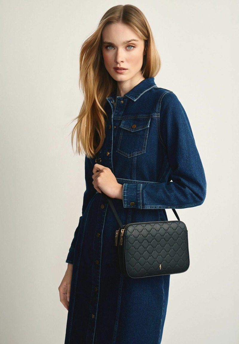 Młoda kobieta z długimi blond włosami, ubrana w ciemnoniebieską sukienkę dżinsową, trzyma czarną pikowaną torebkę na ramię z złotymi detalami zamka.
