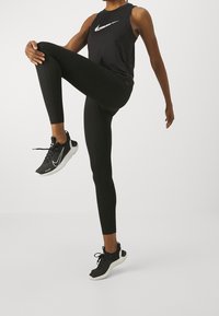 Osoba w czarnym stroju sportowym wykonująca unoszenie kolana w stojącej pozycji, nosząca czarno-białe sneakersy Nike na jednolitym białym tle.