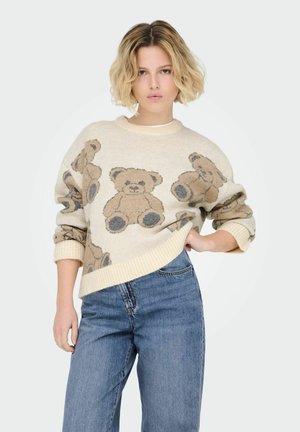 Beige gebreide trui met een teddybeerpatroon in bruin en grijs. Ribgebreide halslijn en manchetten; gecombineerd met lichtblauwe wijde jeans.