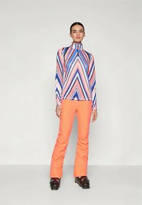 Μακρυμάνικη μπλούζα με φερμουάρ και ζωηρό σχέδιο σε ζιγκ-ζαγκ σε ροζ, μπλε και λευκό, συνδυασμένη με φωτεινές πορτοκαλί παντελόνες και μαύρες μπότες σκι.